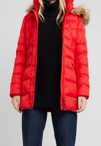 Rød polstret jakke med hætte kantet med pels, lynlåslukning foran og elastiske manchetter. Båret over en sort strikket sweater og mørkeblå jeans.