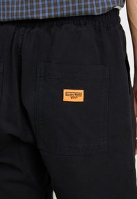 Schwarze Cargo-Hosen mit elastischem Bund, ausgestattet mit zwei Seitentaschen und einem rechteckigen orangefarbenen Etikett mit der Aufschrift "Service Works."