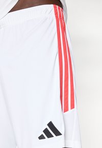 Λευκά αθλητικά σορτς με κόκκινες τρεις ρίγες και μαύρο λογότυπο. Κατασκευασμένα από ελαφρύ ύφασμα με διακοσμητικά δίχτυ κατά μήκος της πλευράς.