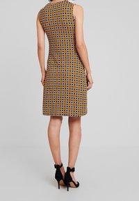 Vestido sin mangas de longitud a la rodilla con un patrón geométrico en amarillo, negro y blanco. Combinado con zapatos de tacón alto negros con correas en el tobillo.