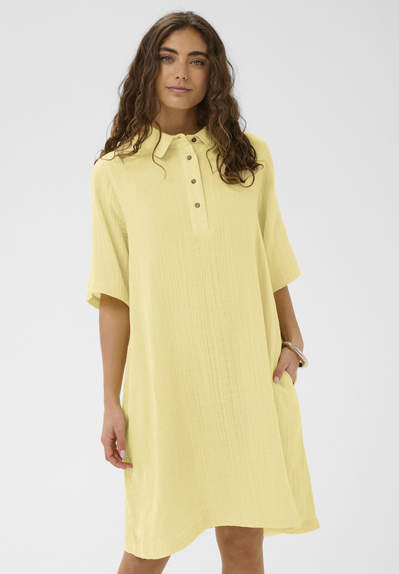 Femme aux longs cheveux bouclés portant une robe jaune ample, boutonnée, arrivant au genou, avec des poches et un bracelet argenté à un poignet.