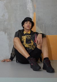 Czarne buty do wędrówek z czerwonymi sznurowadłami, czarna kurtka, koszula w żółtą wzorem, czarne szorty; siedzący oparty o szary mur z pomarańczowymi akcentami.
