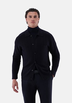 Man standing wearing a black button-up knitted cardigan over a black turtleneck sweater with hands in pockets.