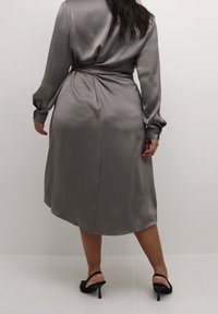 Robe en satin gris à manches longues, taille cintrée et dos drapé. Finition au toucher doux et ourlet légèrement évasé. Sandales noires à talons hauts.