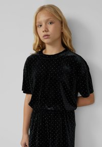 Zwart fluwelen jurk met korte mouwen, voorzien van kleine witte stippen, een elastische tailleband en een ruime pasvorm. Gladde textuur.