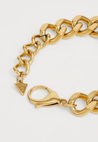 Gouden kettingarmband met gladde, stevige schakels en een driehoekige hanger met "GUESS" gegraveerd. Sluiting met haak inbegrepen.