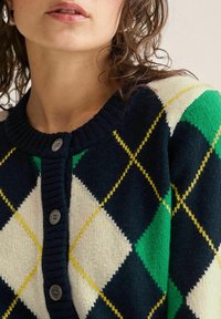 Nainen, joka pukeutuu tummansiniseen, vihreään, valkoiseen ja keltaiseen argyle-kuvioiseen napitettuun neuletakkiin, ja jonka kiharat ruskeat hiukset näkyvät hartioilla.