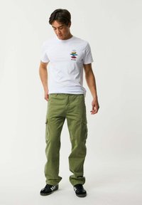 Maglietta bianca in cotone con logo grafico colorato sul petto, abbinata a pantaloni cargo verde oliva a gamba larga e sneaker nere.