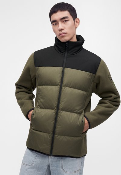 Jeune homme portant une veste matelassée vert olive et noir, les mains dans les poches, debout devant un fond blanc uni.