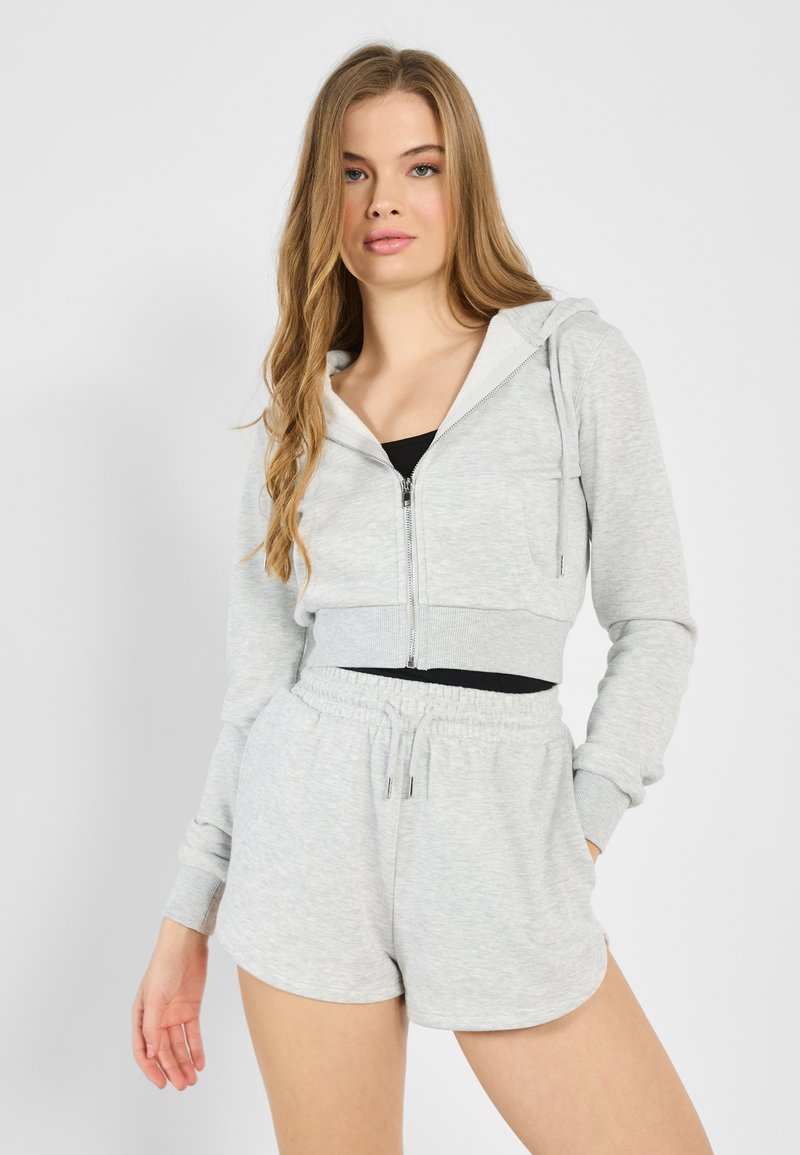 Haut de sweatshirt zippé gris coupé avec des shorts à cordon, le tout en tissu doux et texturé, avec des poches avant et un débardeur noir en dessous.