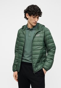 Jachetă verde ușoară, matlasată, cu glugă, prevăzută cu fermoar frontal și un logo pe piept, purtată peste un tricou polo asortat.