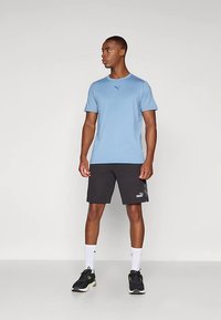Modelo masculino de pie mirando hacia la izquierda, vistiendo una camiseta Puma azul claro, shorts Puma negros, calcetines blancos y zapatillas negras sobre un fondo liso.