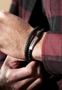 Svart flätad läderarmband med en metallspänne. Bärs på en handled, vilket visar en texturerad yta och en stilren design.