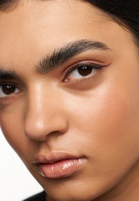 Close-up van een gezicht met gladde, lichtbruine huid, gedefinieerde wenkbrauwen, warme bruine ogen en glanzende lippen. Subtiele make-up accentueert de natuurlijke kenmerken.