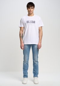 Camiseta blanca de manga corta con "BIG STAR" impreso en letras oscuras, combinada con jeans azules y zapatillas blancas. Diseño sencillo, corte casual.
