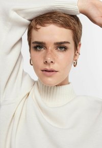 Weißer Strickpullover mit hohem Rippenkragen, getragen von einer Person mit kurzen Haaren und Creolen. Hat einen lockeren Schnitt und ein strukturiertes Gewebe.