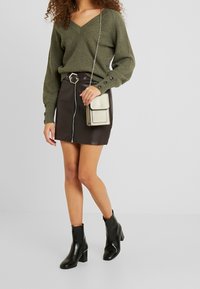 Kobieta ubrana w oliwkowy sweter z dekoltem w serek, czarną skórzaną mini spódniczkę z zamkiem, czarne botki za kostkę i trzymająca małą beżową torbę na ramię.