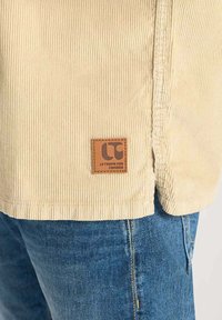 Beige corduroy shirt met gestructureerde stof, voorzien van een bruin leren label met "LC" aan de zoom. Gecombineerd met donkerblauwe denim jeans.