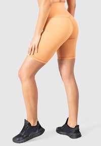 Vysoké cyklistické šortky v broskvové barvě vyrobené z elastického, hladkého materiálu. Nošeny s černými sportovními botami. Ukazují délku nohou a střih.