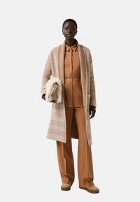 Beige gebreide lange cardigan met gestructureerde strepen, gedragen over een bruine knoop shirt en bijpassende broek. Model houdt een kleine tas met bontaccent.