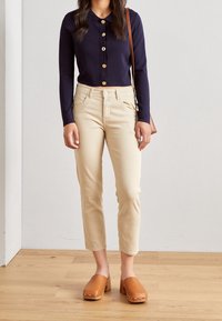 Camisa de manga larga azul marino con botones, pantalones ajustados beige y zuecos marrones. La camisa tiene un collar y botones dorados; los pantalones tienen bolsillos delanteros.