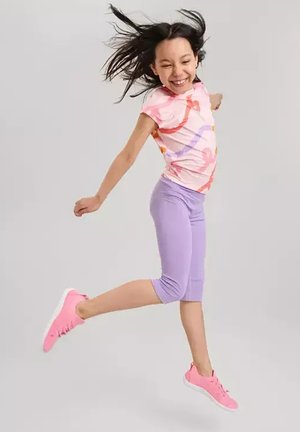 Fille aux longs cheveux noirs portant une chemise rose à motifs et des leggings violets saute joyeusement avec des baskets roses sur un fond clair uni.