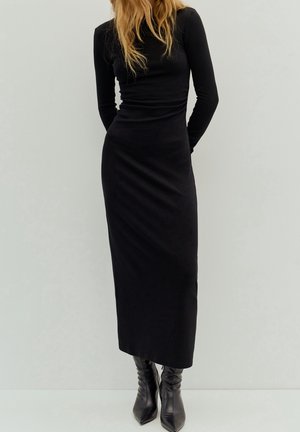 Woman wearing a fitted long-sleeve black midi dress with black pointed-toe boots, standing against a plain light background.