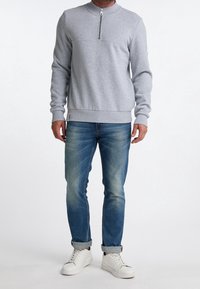 Grauer Pullover mit hohem Kragen und Frontreißverschluss, kombiniert mit blauen Jeans und weißen Sneakers. Glattes Material und lässige Passform.