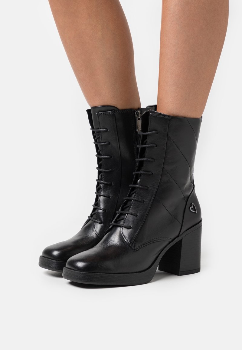 Bottes noires en cuir à lacets avec talons blocs portées sur jambes nues, avec fermetures éclair latérales et un petit détail en forme de cœur près du talon.