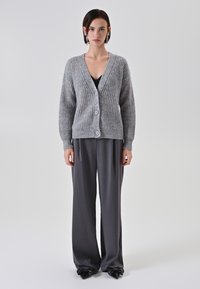 Cardigan grigio a maglia con scollo a V, chiusura con bottoni e texture a costine, indossato sopra una maglietta nera e abbinato a pantaloni grigi a gamba larga.