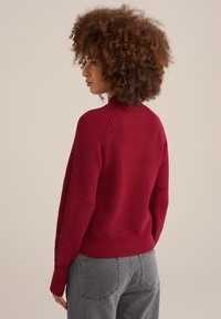 Raudona megzta palaidinė su raukšlėtu tekstūru ir aukštu koloriumi, derinama su pilkais aukštu liemeniu džinsais. Galinis vaizdas rodo formai pritaikytą dizainą.