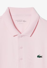 Polo Lacoste rosa claro con cuello texturizado, tres botones y un logo de cocodrilo verde en el pecho izquierdo. Corte regular.