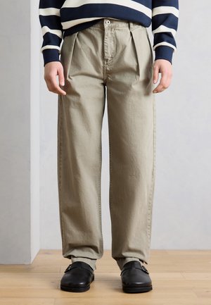 Személy bézs pliszírozott nadrágot, fekete csatos cipőt és sötétkék, fehér vízszintes csíkos pulóvert visel, áll egy fapadlón.