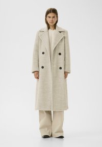 Manteau long à double boutonnage en tissu texturé gris clair avec de larges revers et quatre boutons noirs. Associé à un pantalon beige à jambes larges et des chaussures noires.
