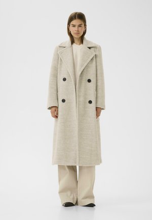Long, double-breasted coat in light gray textured fabric with wide lapels and four black buttons. Paired with beige wide-leg pants and black shoes.