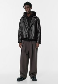 Crna kožna motoristička jakna preko crnog duksera, u kombinaciji s opuštenim sivim hlačama i crnim robustnim cipelama. Glatka tekstura, minimalistički dizajn.