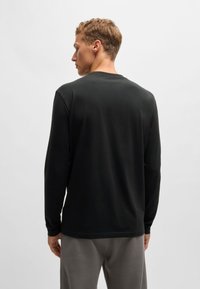 Cămașă neagră cu mâneci lungi dintr-un material din bumbac ribat, având un gât rotunjit și o croială lejeră, asortată cu pantaloni gri.