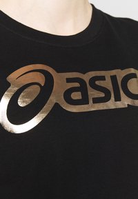 Camiseta negra con cuello redondo que presenta un gran logo "Asics" en dorado metálico, destacado en el centro del pecho. Tela de textura suave.