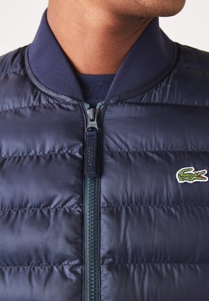 Chaqueta acolchada azul marino con cierre de cremallera, cuello acanalado y logo de cocodrilo bordado en verde en el pecho. Presenta una textura acolchada.