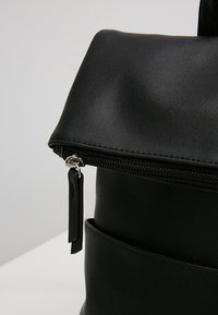 Černý batoh z umělé kůže s přehazovacím klopou, zipovým zapínáním a kovovými detaily. Vyznačuje se hladkou texturou a minimalistickým designem.