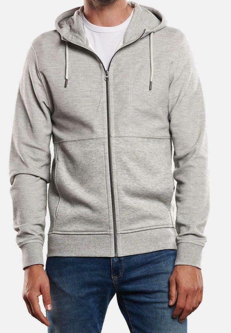 Homme portant un sweat à capuche gris clair zippé par-dessus un t-shirt blanc et un jean bleu, debout les bras détendus le long du corps.