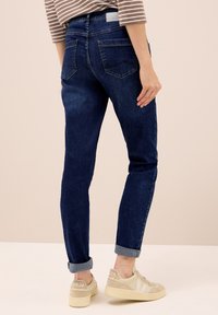 Jeans de denim azul oscuro con un corte ajustado, que cuentan con cinco bolsillos y bajos acampanados, combinados con zapatillas beige. Textura suave y diseño clásico.