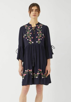 Mörkblå klänning med blommig broderi i rosa och lila. Har en nyckelhålsringning, klockärmar och en lös, flödande silhuett.