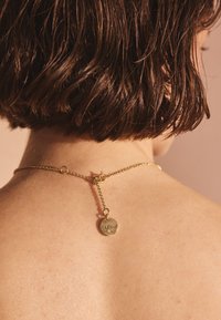 Kort brunt hår ovanför axlarna, guldkedjehalsband fastsatt i nacken med ett runt hängsmycke ingraverat med "Carla Kubera."