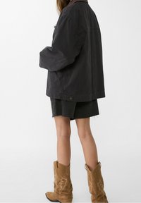 Persoon in een oversized donker jack, versleten zwarte shorts en beige suède cowboylaarzen staat tegen een effen witte achtergrond.