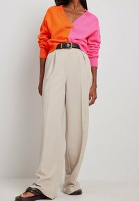 Pull tricoté bicolore en orange et rose avec un col en V, associé à un pantalon beige plissé taille haute et des sandales marron à bout ouvert.