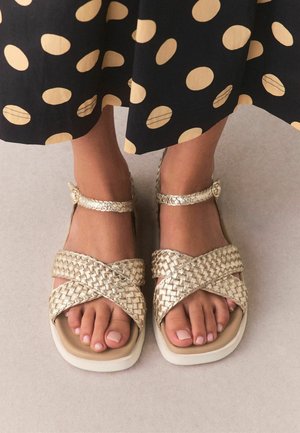 Feet wearing woven gold cross-strap sandals with ankle straps, paired with a black skirt featuring large beige polka dots.