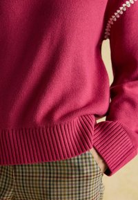 Pulover tricotat fuchsia cu tiv striat, având accente decorative albe pe mâneci, purtat peste pantaloni în carouri în tonuri atenuate.