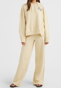 Felpa beige con logo frontale e pantaloni a gamba larga coordinati. Entrambi sono realizzati in un tessuto morbido e liscio, con un fit rilassato e polsini a costine.