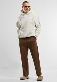 Light gray hoodie with a front pocket and blue text, paired with brown, pleated trousers and tan slip-on shoes. Accessories include sunglasses.